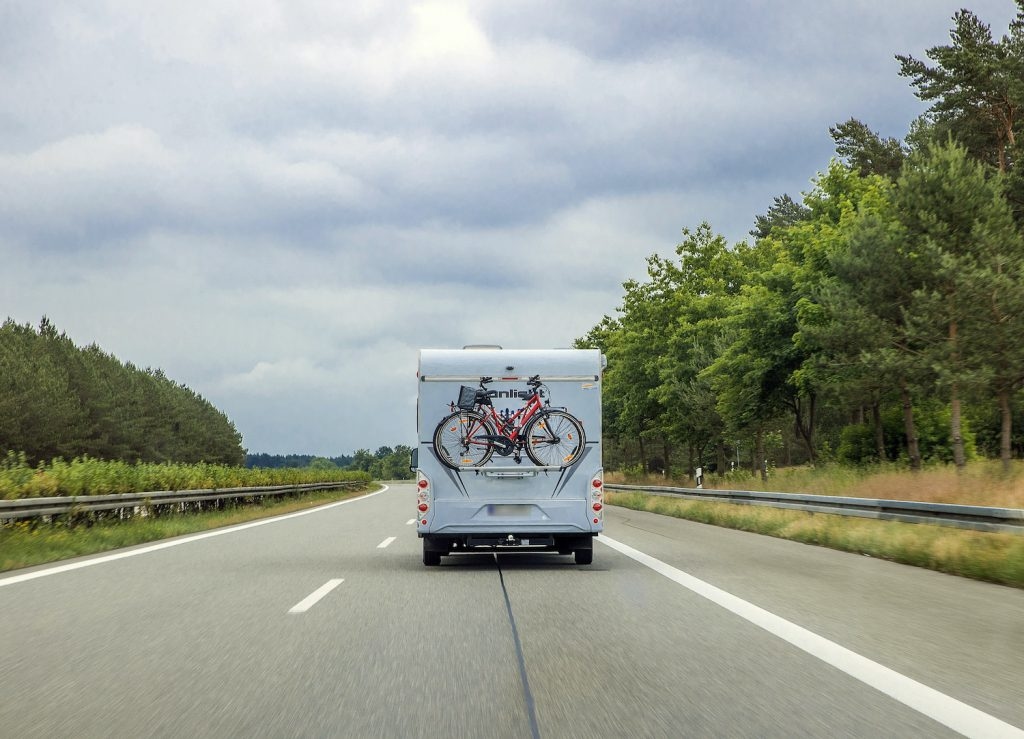 A class C RV driving down the road. Only the back of the RV is visible. There is a red bike mounted o the back. 