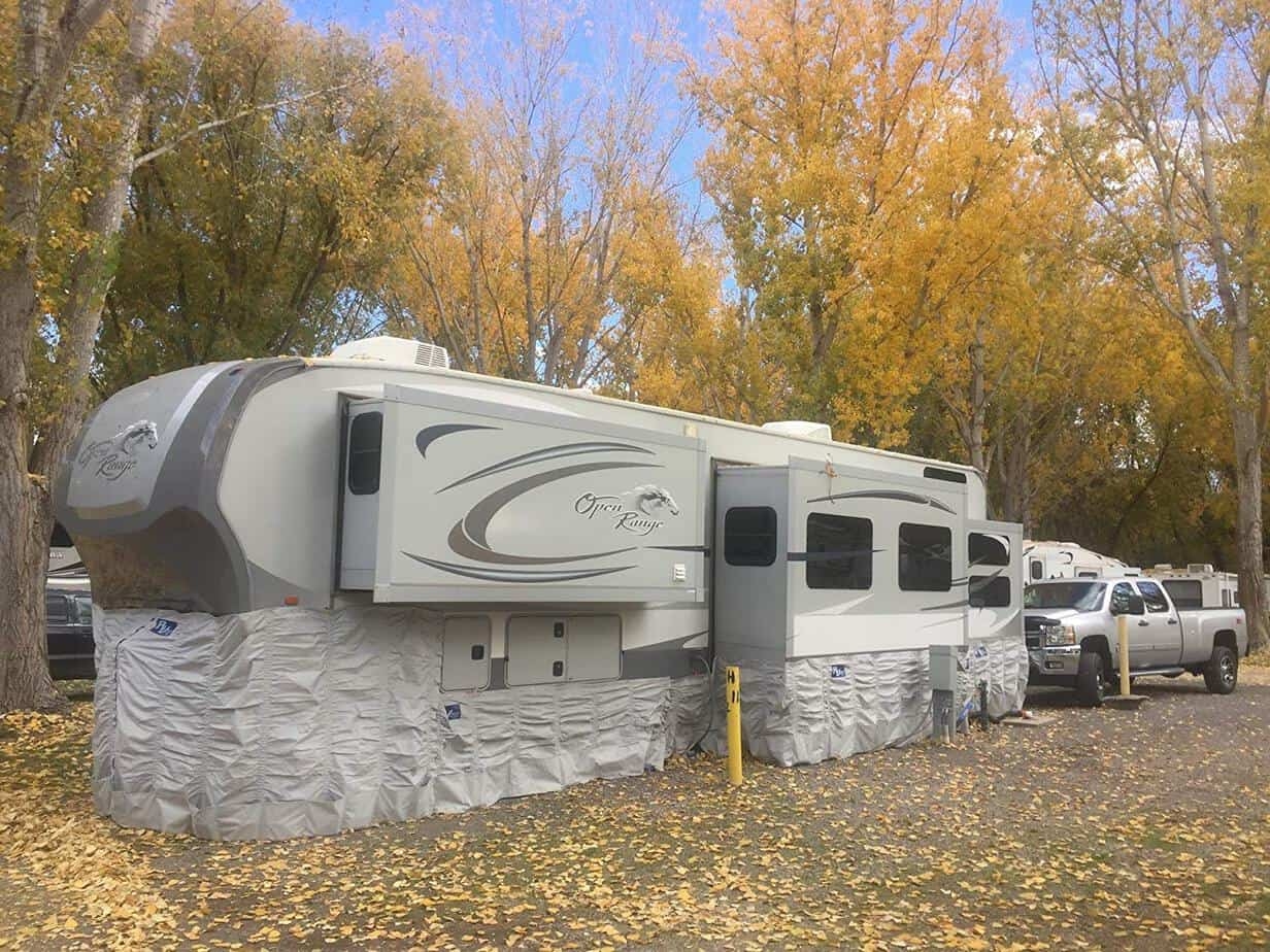 A fifth wheel RV stationary at an RV park in the fall. There is insulated skirting wrapped around the entire bottom of the trailer to help prevent frozen pipes. 